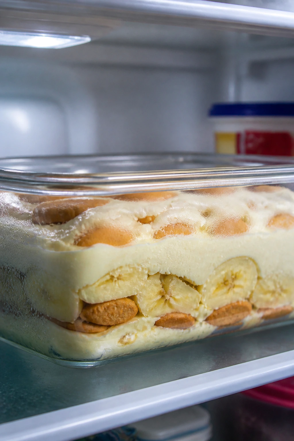 Banana pudding in a glass container inside a fridge with condensation on the glass